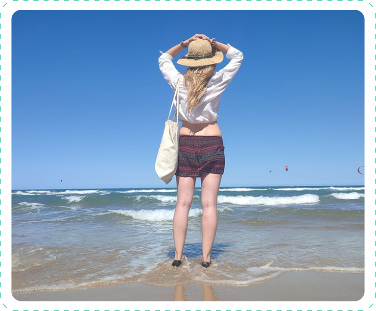 A woman stands on the shore, facing the sea with hands on her hat.